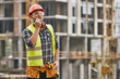 © Friends Stock - Сhecking work. Professional young builder in working uniform and red protective helmet talking to crane operator by walkie talkie while standing at construction site.