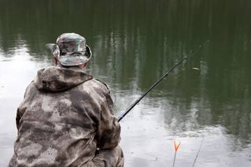 Fisherman in camouflage clothes sitting with a fishing rod on the shore. Man angling on the lake in spring, trees are reflected in the water