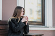 © spaskov - Young beautiful millenial girl in a coat is sitting on an outdor bench with a paper cup of coffee. Autumn or spring day, cool weather.
