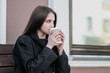 © spaskov - Young beautiful millenial girl in a coat is sitting on an outdor bench with a paper cup of coffee. Autumn or spring day, cool weather.