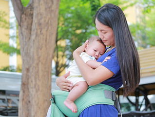 Beautiful Asian mother hugging her infant on ergonomic baby carrier in the garden.