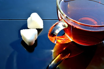 Glass Cup of brown black hot sweet tea and two pieces of sugar in shape of heart with reflection on black metal background in sun light