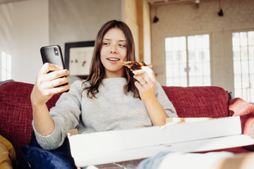 Beautiful girl eating delivery pizza from the box and taking selfie photo using mobile phone camera