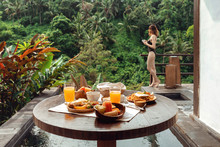 Beautiful Breakfast On A Wooden Table Amid The Pool With A Young Woman Holding A Cup Of Coffee In Hand. Sumptuous Breakfast At A Hotel Overlooking The Tropical Trees, Ubud, Bali