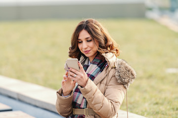 Caucasian woman in jacket and with scarf sitting on the bench outside and writing or reading message on smart phone.