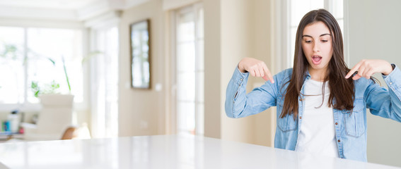 Wide angle picture of beautiful young woman sitting on white table at home Pointing down with fingers showing advertisement, surprised face and open mouth