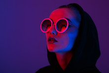 Portrait Of Fashion Young Girl In A Black Sweater With A Hood And Round Sunglasses In Red And Blue Neon Light In The Studio