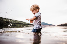 A Wet, Small Toddler Boy Standing Outdoors In A River In Summer, Playing.