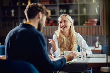 Smiling Caucasian blonde businesswoman dressed smart casual discussing with her male colleague about project while sitting in coffe shop.