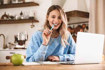 Portrait of attractive blond woman 20s wearing casual sweatshirt working on laptop and writing down notes at home