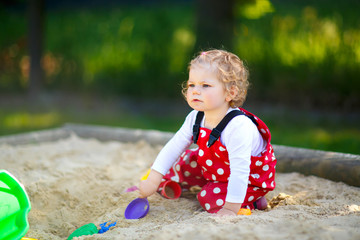 Cute toddler girl playing in sand on outdoor playground. Beautiful baby in red gum trousers having fun on sunny warm summer day. Child with colorful sand toys. Healthy active baby outdoors plays games