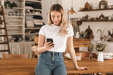 Poster - Portrait of charming blond woman using smartphone while standing in stylish wooden kitchen at home