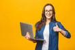Portrait of a smiling young beautiful girl holding laptop computer and showing thumbs up isolated over yellow background.