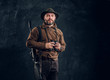 Mature hunter with rifle holding binoculars and looking at a camera. Studio photo against dark wall background