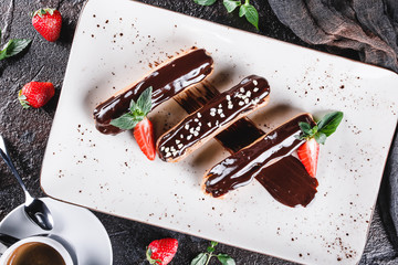 Wall Mural - Homemade cake eclairs or profiteroles with custard, chocolate and strawberries on dark background served with cup of coffee. Traditional french eclairs, dessert. Top view, flat lay