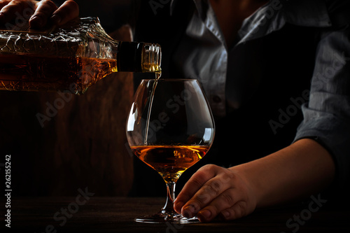 The bartender pours the cognac or brandy in big wine glass on the old bar counter. Vintage wooden background in pub or bar, night mood. Place for text, toning, selective focus