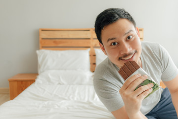 Man is eating chocolate bar on the bed in his bedroom.