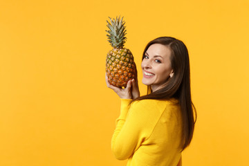 Smiling young woman in casual clothes looking back, holding fresh ripe pineapple fruit isolated on yellow orange wall background. People vivid lifestyle, relax vacation concept. Mock up copy space.