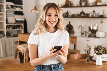 Sticker - Portrait of modern blond woman using mobile phone while standing in stylish wooden kitchen at home