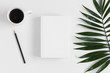 Top view of a white book mockup with  workspace accessories and a palm leaf on a white table.