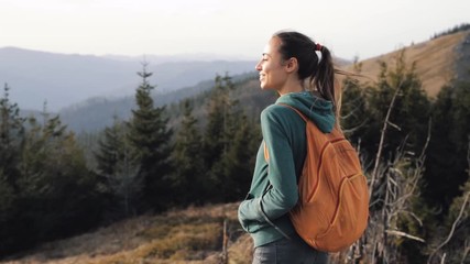 Wall Mural - pretty woman hiker with small orange backpack is standing on edge of mountain ridge. Woman is hiking in mountains before sunset. strong wind is fluttering woman's hair