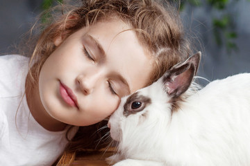 Wall Mural - Little girl playing with real rabbit. Child and white bunny on Easter on flower background. Kid kiss pet. Fun and friendship for animals and children.