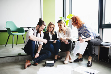 Wall Mural - A group of young business people sitting on the floor in an office, talking.