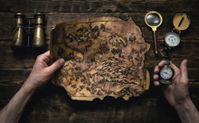 Old Map In Man Adventurer Hands On A Brown Table Background. Treasure Hunt Concept.