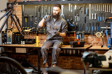 Portrait of a handsome repairman in workwear sitting with wrenches and different work tools at the bicycle workshop