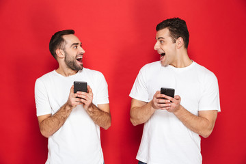 Wall Mural - Image of two optimistic men friends 30s in white t-shirts smiling and holding smartphones together