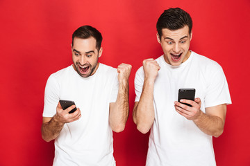 Wall Mural - Image of two caucasian men friends 30s in white t-shirts smiling and holding smartphones together