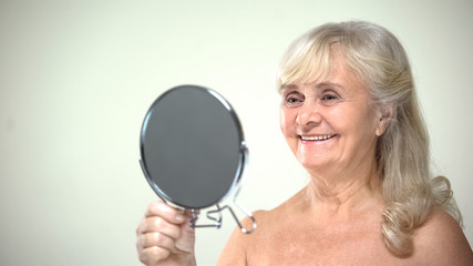 Wall Mural - Positive woman looking in mirror, satisfied with cosmetic procedures, skincare