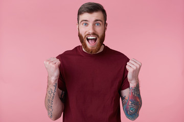 Close up of bearded young man with tattooed hand, screaming and cheering for his favorite team, looking at camera isolated over pink background. People and emotion concept.