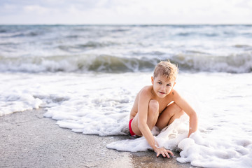 Little cute caucasian toddler boy with blond hair plays at the beach in sea waves on sunny summer day. Family vacation with kids fun happiness closeness unity with nature. Authentic lifestyle