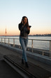 © dissx - Young woman listens to music in closed headphones through her phone wearing a leather jacket and jeans at a sunset near river Daugava