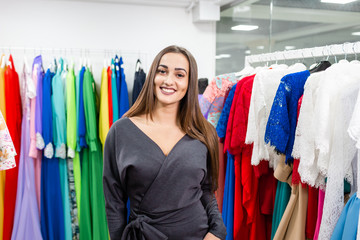 Portrait of a girl on the background of clothes on hangers in the clothing store. Happy young woman choosing clothes. Sale, fashion, consumerism concept