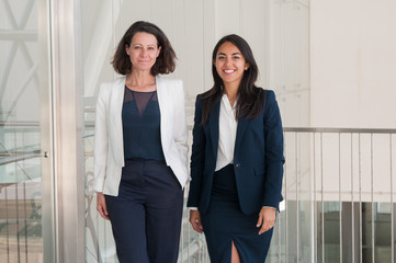 Two positive business women posing at camera in office hall