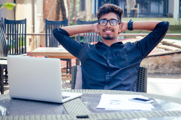 handsome and successful indian man in a stylish well-dressed freelancer working with a laptop on the beach.freelance and remote work.businessman student in a summer cafe on the shore of india ocean