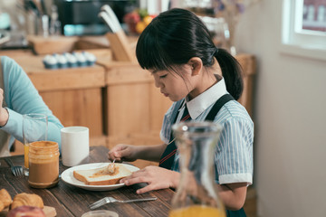 Wall Mural - focus on smart cute little girl having breakfast wear uniform before school in morning. daughter sitting beside mom putting peanut jam on bread toast. family time healthy lifestyle early day.