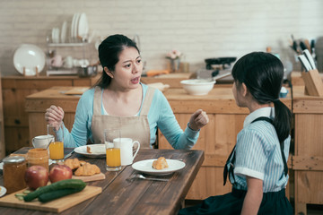 Wall Mural - lazy sleepy little girl wake up late for school sitting at kitchen table with breakfast at home. frowning angry mom loudly nag at daughter bad habit in early morning. unhappy asian mother worried.