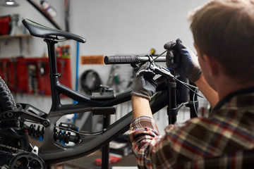 Cropped shot back view of professional mechanic working in bicycle repair shop, man repairing modern bike using special tool, wearing protective workwear and gloves