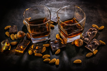 Glasses of whiskey, chocolate with hazelnuts, candy and almonds on a dark wooden background.