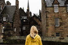 Blonde Woman Backwards Wearing A Yellow Raincoat Looking To A Medieval Housing