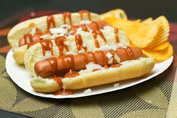 Two New York style hot dogs with ketchup, mustard, onions, and a side of potato chips, served on a paper plate