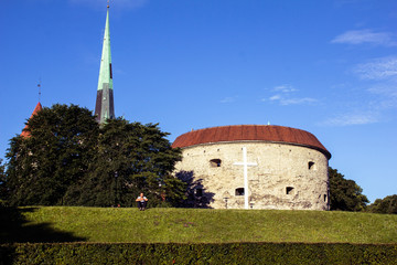 Towers and round building with white cross in front of it in the beginning of old town of Tallinn.  Green alley in front of old town's buildings with the man sitting on it with green tree behind him.