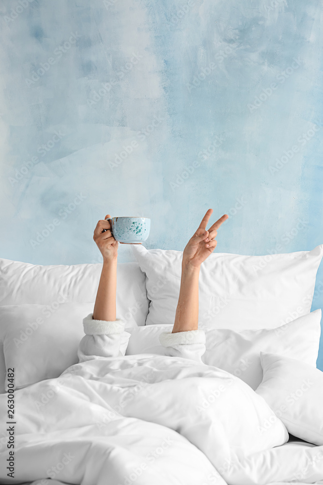 Young woman with cup of hot beverage showing victory gesture in bed - obrazy, fototapety, plakaty 