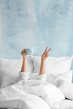 Fototapeta  - Young woman with cup of hot beverage showing victory gesture in bed