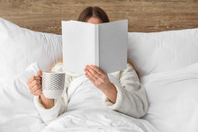 Young woman with cup of hot beverage reading book in bed