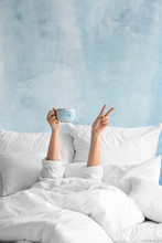 Young Woman With Cup Of Hot Beverage Showing Victory Gesture In Bed