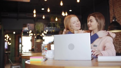 Wall Mural - Tracking right shot of two young women greeting each other with embrace at table in cafe and discussing project on laptop computer during coffee break